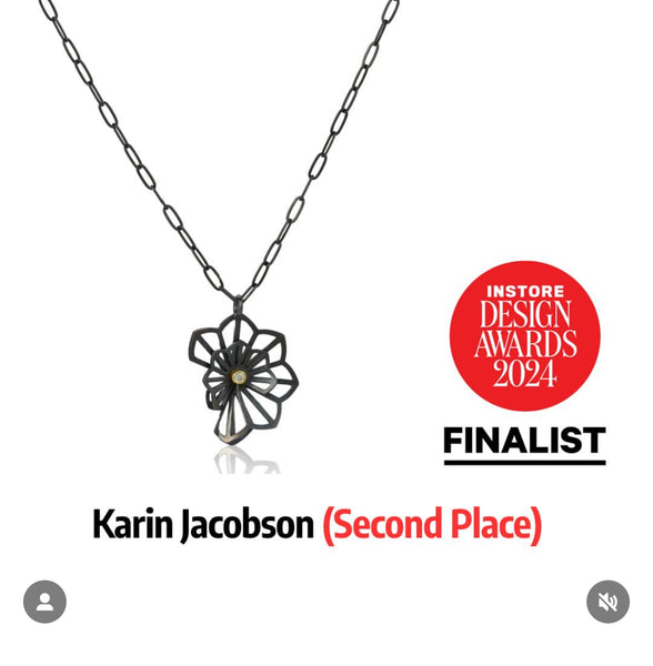 Photo of a pendant in Karin Jacobson's Origami flower style - made in oxidized sterling silver with a diamond set in 18k gold in the middle, shown on a white background. Includes the text "In Store Design Awards Finalist. Karin Jacobson Second Place."